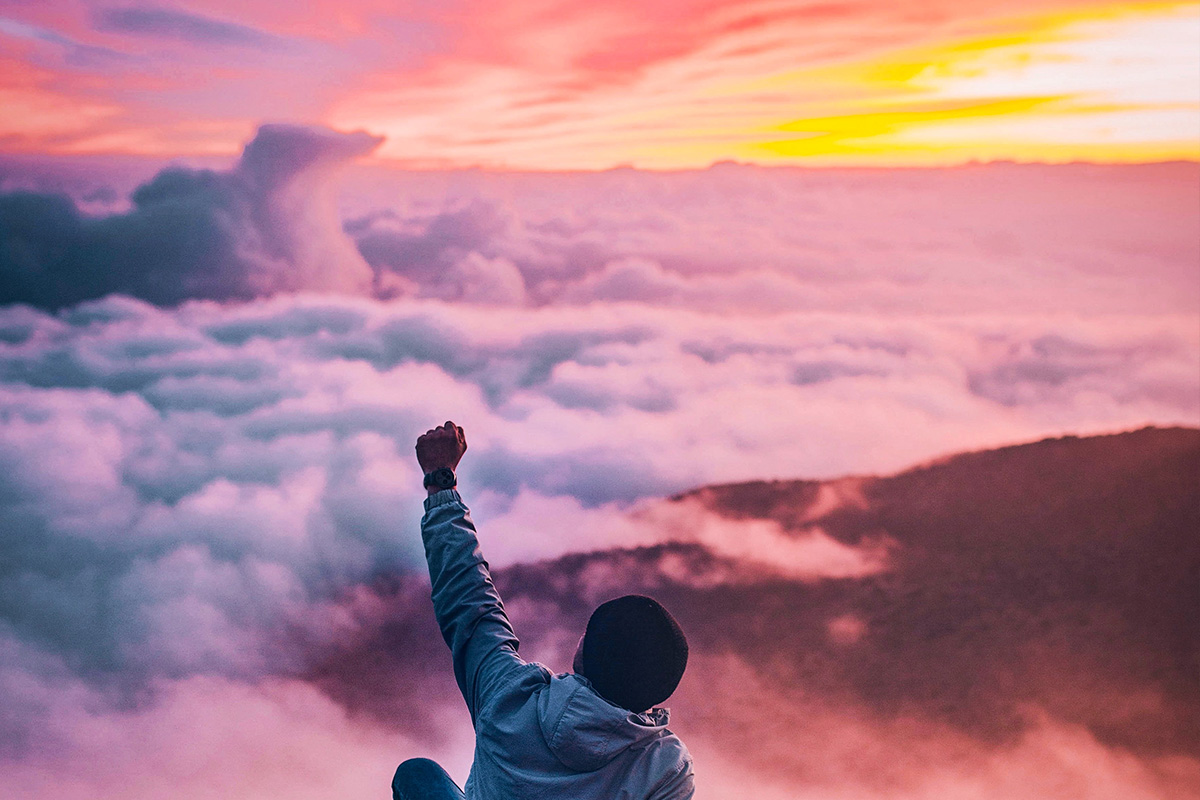 A man in the clouds on top of a mountain. The sun is rising and he has his fist in the air victoriously