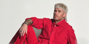 Colton Dixon sitting with his arm resting on his knee., He is wearing a bright red jumpsuit